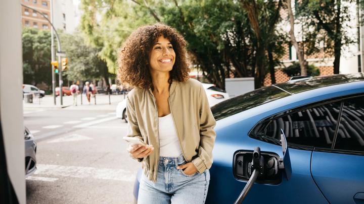 A woman is smiling as she stands next to her electric car at a charging station in a city environment. The scene captures modern transportation and eco-friendly living.