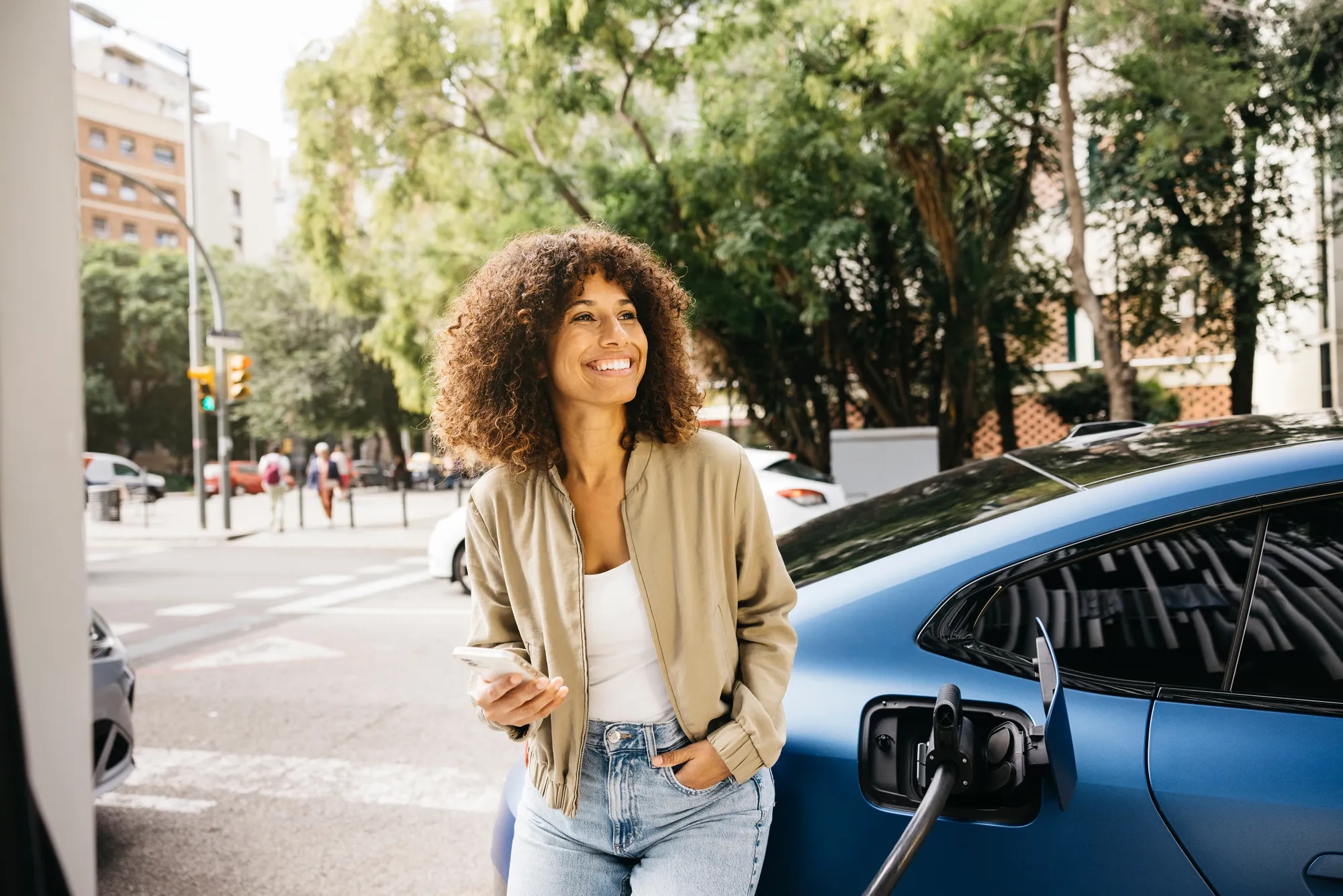 A woman is smiling as she stands next to her electric car at a charging station in a city environment. The scene captures modern transportation and eco-friendly living.
