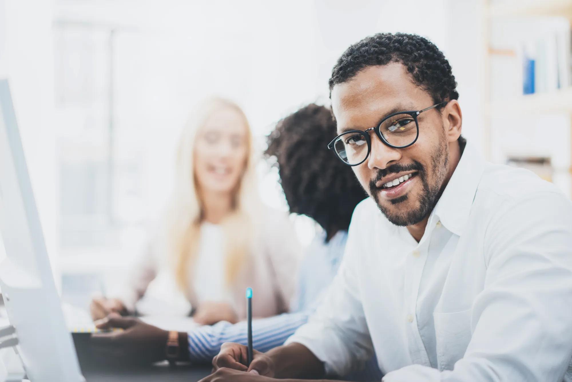 Dark skinned entrepreneur wearing glasses, working in modern office.African american man in white shirt looking and smiling at the camera.Horizontal,blurred background.