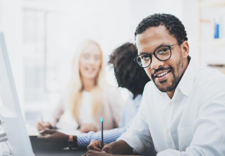 Dark skinned entrepreneur wearing glasses, working in modern office.African american man in white shirt looking and smiling at the camera.Horizontal,blurred background.