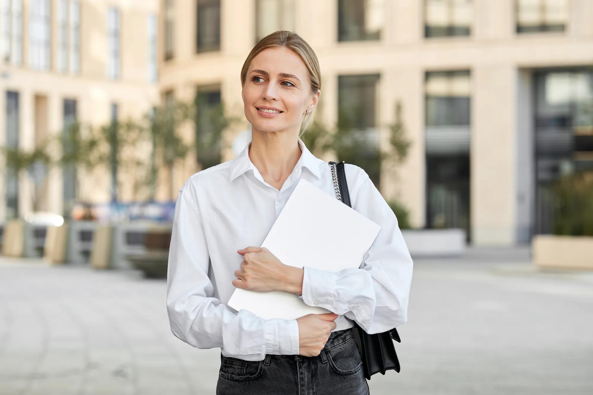 A pretty young woman with white hair dressed in a white shirt and black jeans carries a laptop with her.