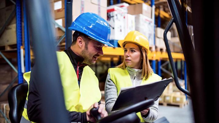 Young warehouse workers working together. Man sitting in a forklift and woman holding notes, discussing something.
