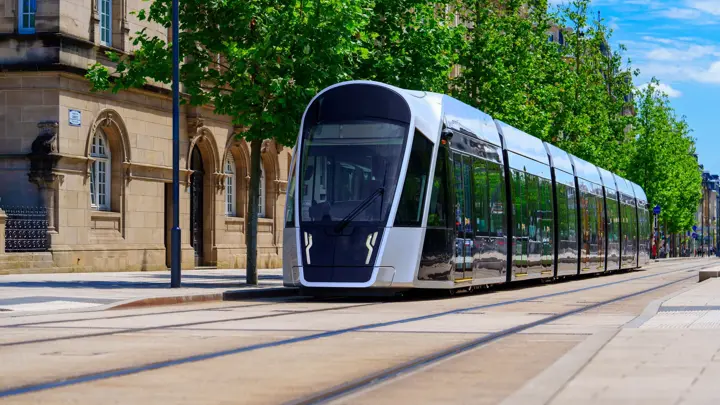 Luxembourg, A modern silver tram moves swiftly along the street, creating a motion blur, while a vintage stone building with many windows stands alongside, surrounded by trees and urban scenery.