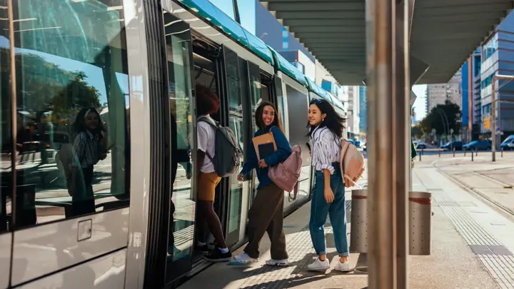 A couple of young stylish students are on their way to school. They are at the train station boarding a train to go to school.