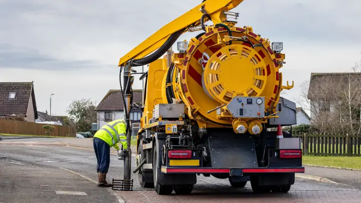 A municipal Medium Volume Combination (MVC) tanker truck operative cleans gully drain removing blockages with suction pipe tool at side of a suburban street. The male operative inserts the metal pipe into the drain.