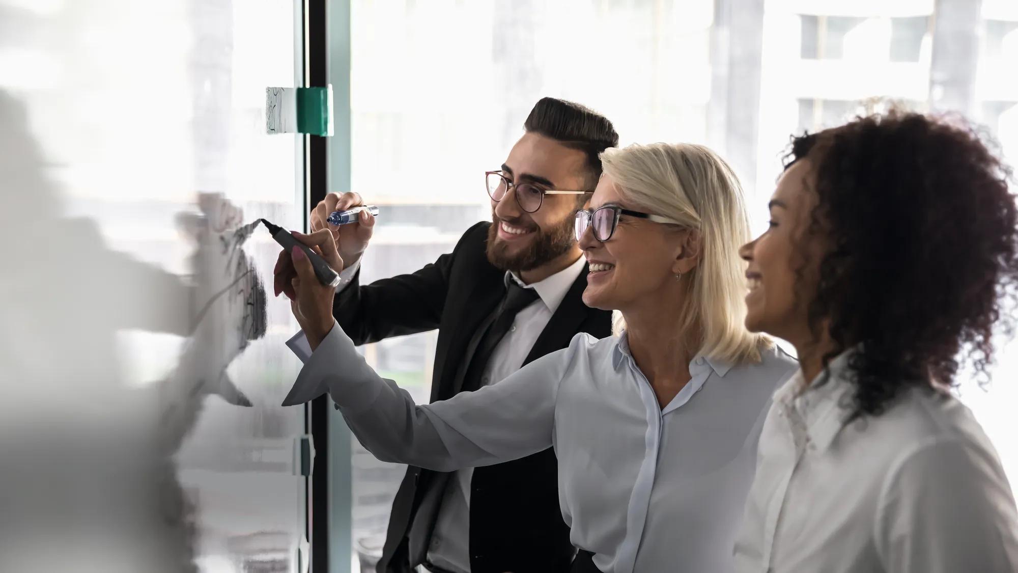Smiling multiethnic team work together write on whiteboard brainstorming discussing new ideas together, happy motivated employees engaged in creative thinking, develop strategy on flip chart
