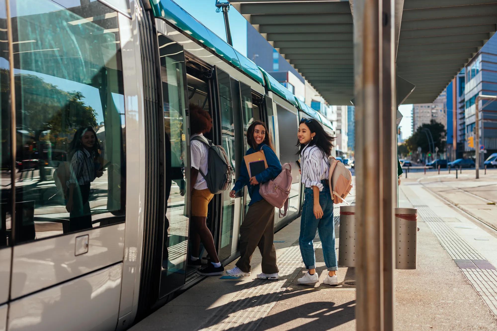 A couple of young stylish students are on their way to school. They are at the train station boarding a train to go to school.