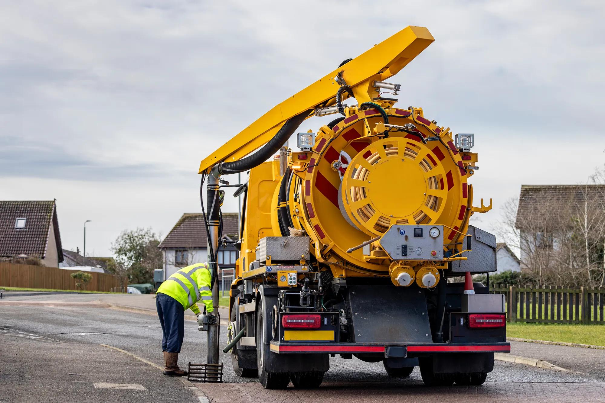 A municipal Medium Volume Combination (MVC) tanker truck operative cleans gully drain removing blockages with suction pipe tool at side of a suburban street. The male operative inserts the metal pipe into the drain.